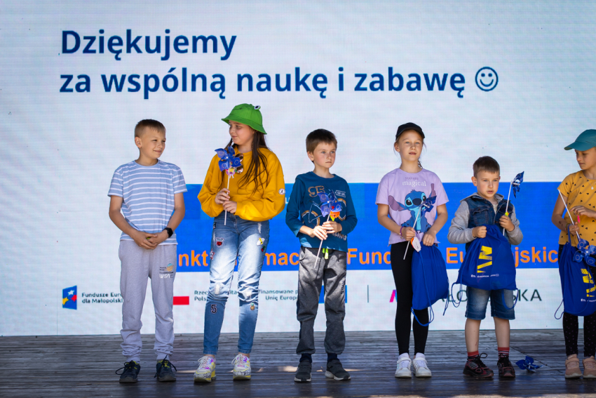 Grupka 6 dzieci w wieku wczesnoszkolnym stojących obok siebie na scenie. Niektóre z nich trzymają w rękach niebieskie wiatraczki z żółtymi gwiazdkami (Znak Unii Europejskiej). Za dziećmi w górnej części zdjęcia napis "Dziękujemy za wspólną naukę i zabawę". Na dole widoczne fragmenty informacji o Funduszach Europejskich.