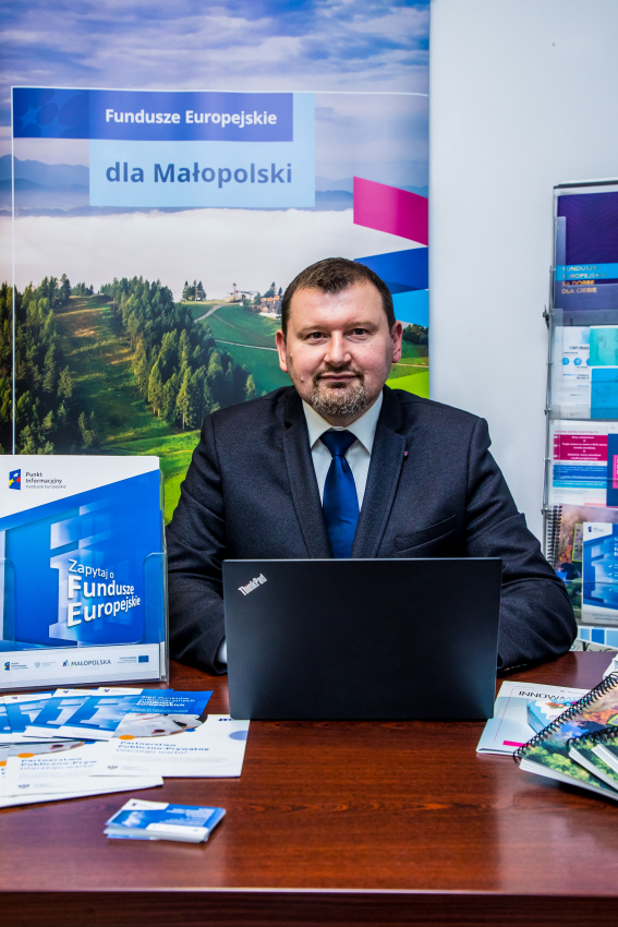 Konsultant Punktu Informacyjnego siedzi przy biurku. Przed nim otwarty laptop. Na biurku leżą ulotki i materiały informacyjne dotyczące Funduszy Europejskich.Za nim plakat z napisem Fundusze Euroepejskie dla Małopolski.