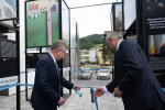 Dwóch starszych, elegancko ubranych mężczyzn rozwiązuje wstęgę na znak otwarcia wystawy. Po obu stronach widoczne metalowe, wysokie, czarne konstrukcje , na których zamieszczone są zdjęcia miejsc w Małopolsce dofinansowanych ze środków Unii Europejskiej. W oddali budynki mieszkalne, zaparkowane samochody, las. 