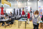Stoisko krawieckie. Stoliki, na których leżą magazyny o modzie i projekty ubrań. Scena, na której stoją manekiny ubrane w eleganckie suknie. Przy stolikach kilka osób.