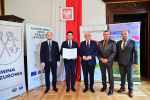 Pięciu mężczyzn w garniturach stoi w eleganckiej sali . jeden z znich trzyma dokument-promesę.