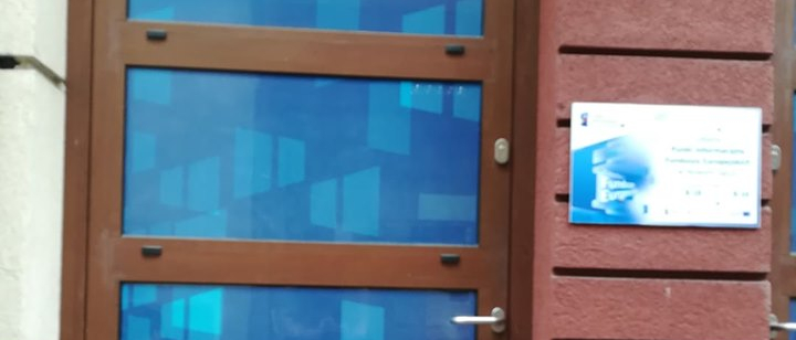 Wejście do budynku. Obok drzwi tabliczka informacyjna.