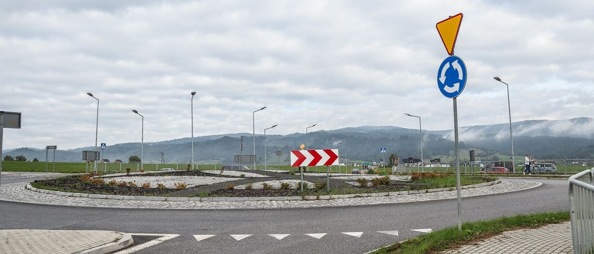 Rondo. Znaki drogowe: znak ustąp pierwszeństwa, znak ronda. W oddali widoczne latarnie, wzgórza, las, zaparkowane samochody i postać. 