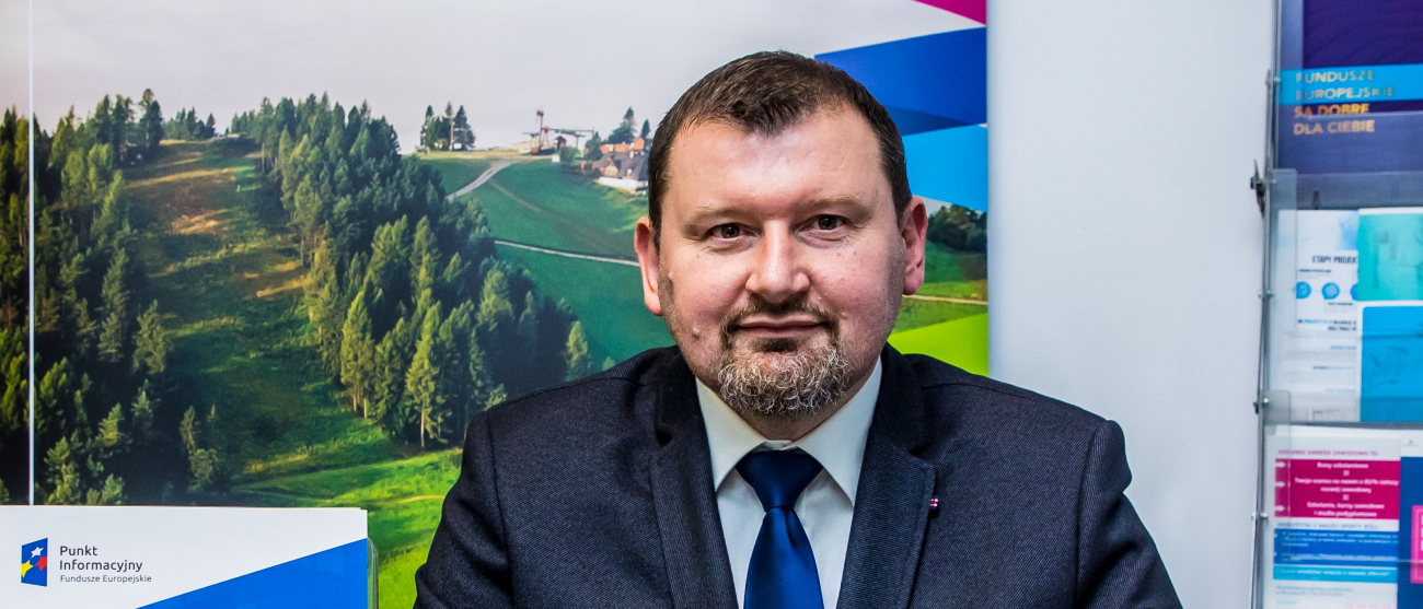 Konsultant Punktu Informacyjnego siedzi przy biurku. Przed nim otwarty laptop. Na biurku leżą ulotki i materiały informacyjne dotyczące Funduszy Europejskich.Za nim plakat z napisem Fundusze Euroepejskie dla Małopolski.