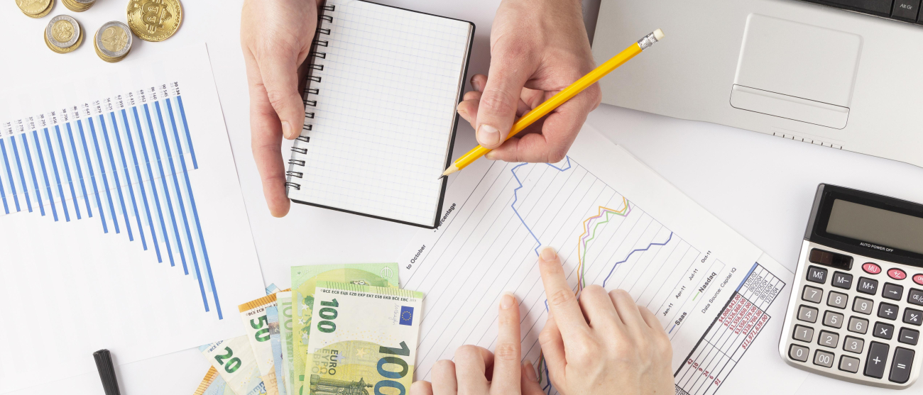 Widoczne dłonie dwóch osób na biurku. Rozłożone dokumenty, wykresy, monety, banknoty euro, kalkulator, okulary, fragment tabletu.